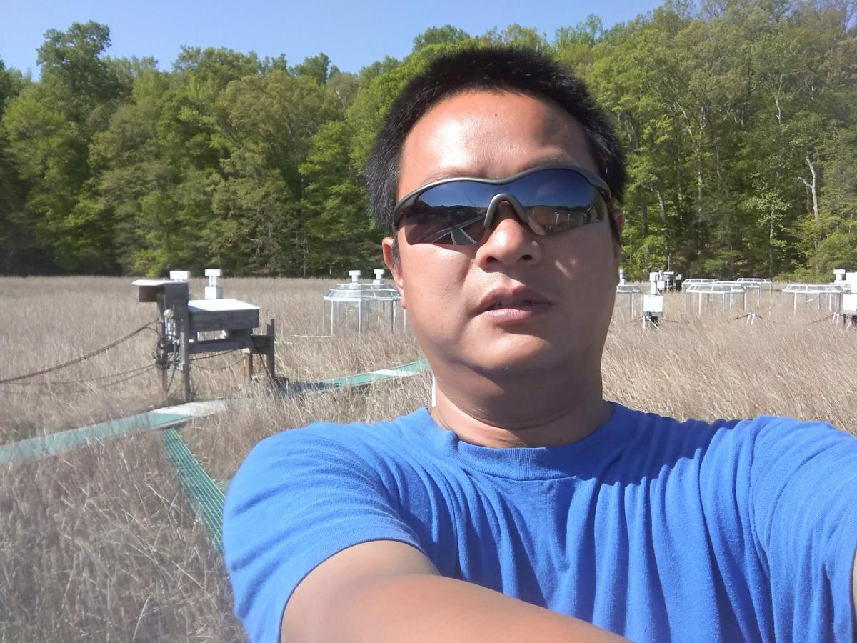 Selfie of a scientist in sunglasses and a blue T-shirt, in front of a wetland with a green boardwalk and experimental chambers